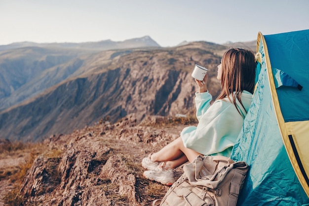 La niña realizó una caminata de montaña activa con una tienda de campaña. Sentada en el suelo con una taza de té en las manos y mirando las montañas rocosas disfrutando de la vista.