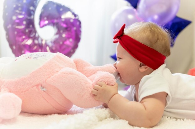 Una niña recién nacida con una falda roja y un lazo rojo en la cabeza está acostada en la cama junto a un juguete de peluche.