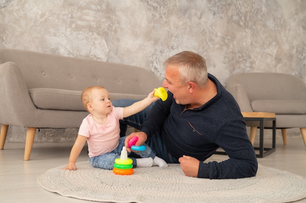 Niña recoge una pirámide con abuelos en la sala de estar. El abuelo juega con la nieta en el suelo cerca del sofá. El niño le da un pato de juguete al abuelo.