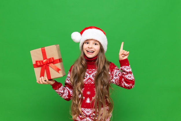 Una niña con un regalo en la mano con un sombrero de Papá Noel sonriendo ampliamente y señalando con el dedo índice hacia arriba sobre un fondo verde aislado Regalos de Navidad para niños