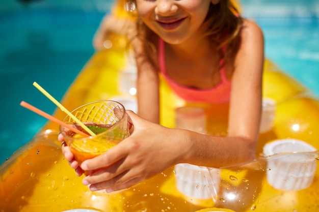 Niña relajándose en la piscina, disfrutando del bronceado, bebe un jugo en un colchón amarillo inflable en el agua en vacaciones familiares