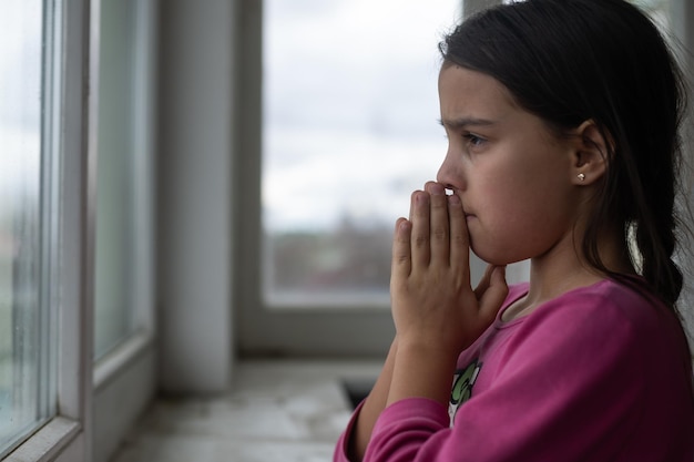 Niña rezando cerca de la ventana.