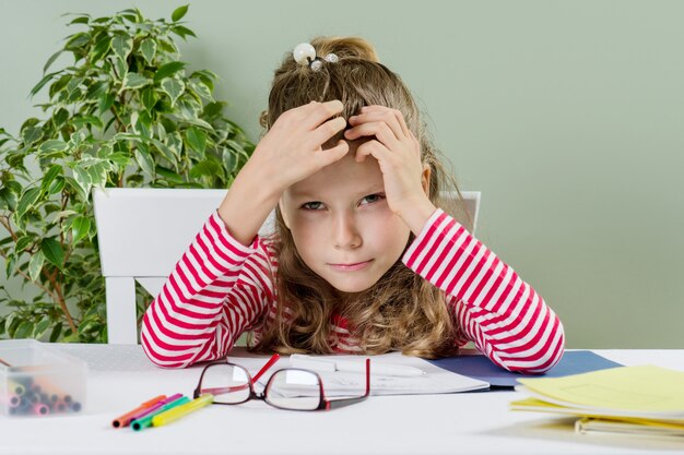 Foto niña ríe, se sienta en un escritorio, dibuja y escribe, las manos con la cabeza.