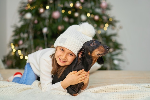 Una niña se ríe con su amigo, un perro salchicha, cerca del árbol de Navidad.