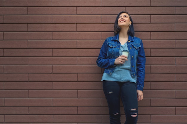 Niña riendo en chaqueta de jeans cerca de una pared de ladrillo marrón con una taza de café en la mano mirando hacia arriba.