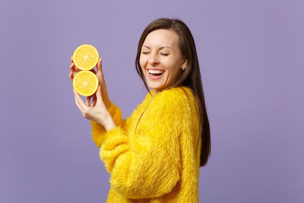 Niña riéndose con suéter de piel manteniendo los ojos cerrados sosteniendo mitades de fruta fresca de naranja madura aislada en un fondo violeta pastel. El estilo de vida vívido de la gente relaja el concepto de vacaciones. Simulacros de espacio de copia.