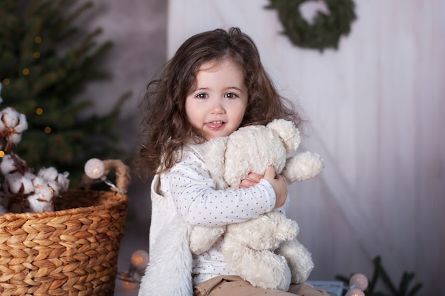 Niña rizada abraza al oso de peluche cerca del árbol de Navidad.