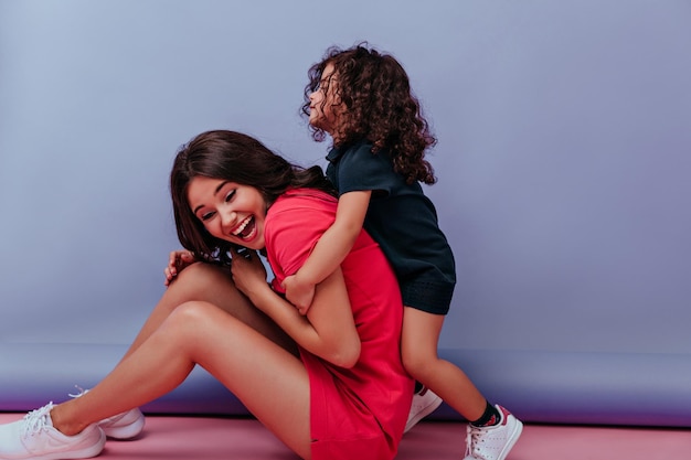 Niña rizada jugando con su madre con fondo morado Foto interior de una familia dichosa jugando durante la sesión de fotos