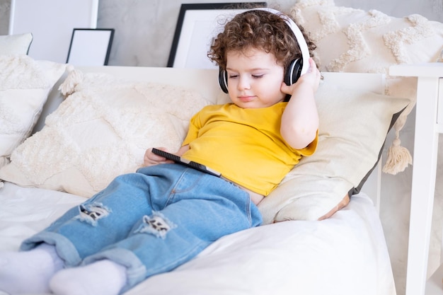 Niña rizada de niño feliz sonriente usando teléfono en auriculares inalámbricos en la cama en casa