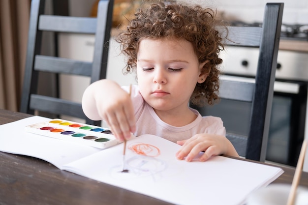 Niña rizada pintando con pinturas de acuarela en casa en la mesa de la cocina