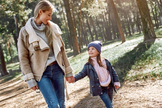 Una niña rizada con sombrero está caminando con su madre en el bosque. Temporada fría, el sol brillante se ve a través de los árboles. De cerca