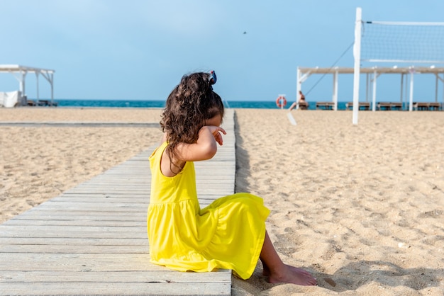 Niña rizada en un vestido amarillo se sienta en una playa desierta de arena en un día soleado de vacaciones divertidas