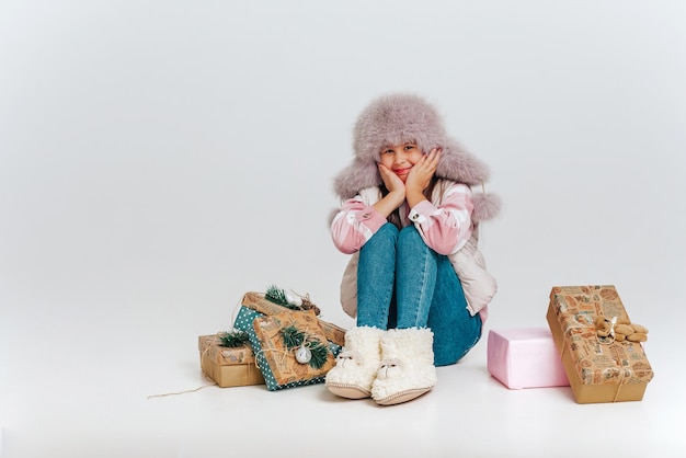 Una niña en ropa de abrigo ang con un sombrero de piel está sentada sobre un fondo blanco rodeado de regalos Espacio