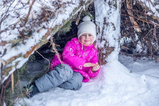 Niña en ropa de abrigo juega en una cabaña de ramas de coníferas en el bosque de invierno, pasar tiempo al aire libre en invierno