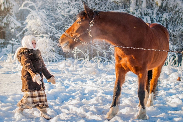 Niña con ropa antigua se estira sorprendida al ver un caballo en un día soleado de invierno helado.