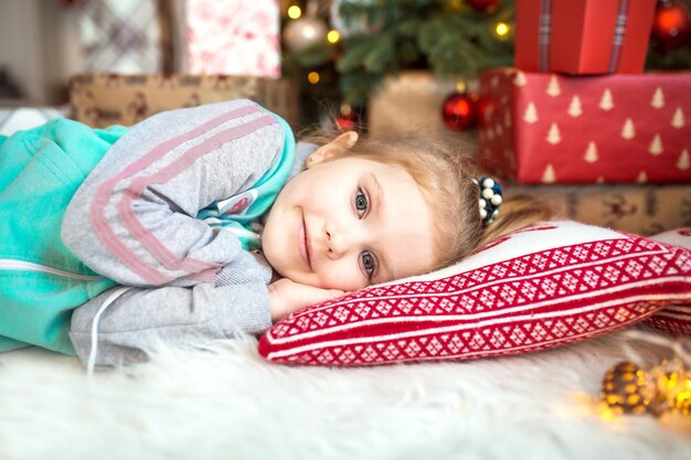 Una niña con ropa de casa yace bajo el árbol de Navidad cerca de las cajas con regalos.