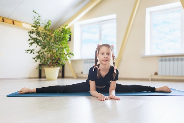 Niña en ropa deportiva negra practicando yoga realiza una división transversal sobre una colchoneta de gimnasia el ejercicio Samokanasana