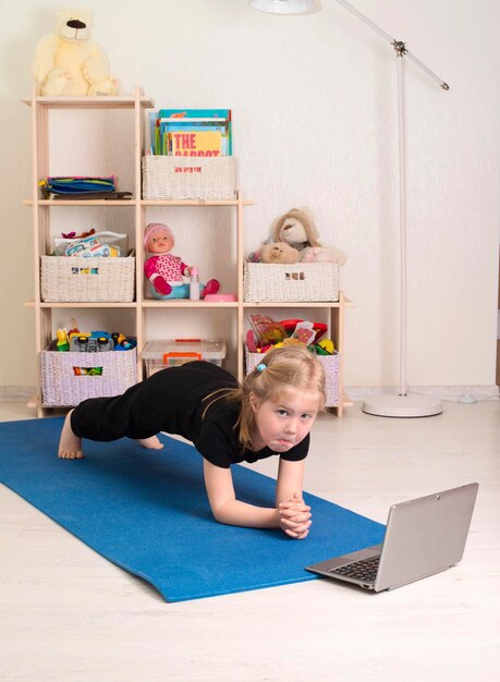 Niña en ropa deportiva viendo videos en línea en una computadora portátil haciendo ejercicios de fitness en casa