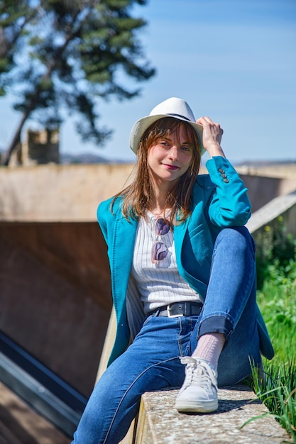 Niña con ropa informal y sombrero blanco sentada en un banco mirando a la cámara en un día soleado