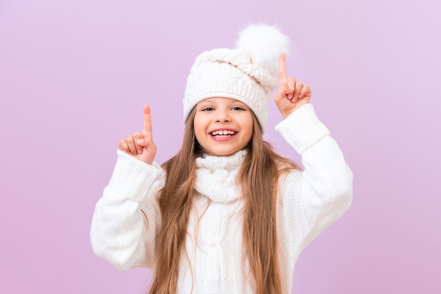 Una niña en ropa de invierno señala su anuncio sobre un fondo rosa aislado.