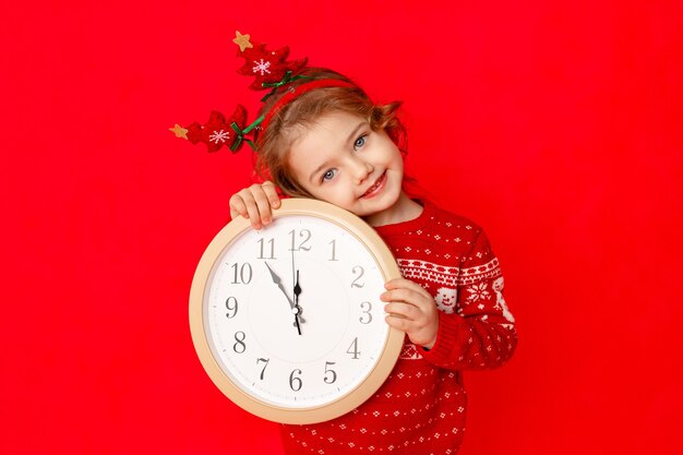 Una niña en ropa de invierno sostiene un reloj sobre un fondo rojo. Concepto de año nuevo, espacio para texto.