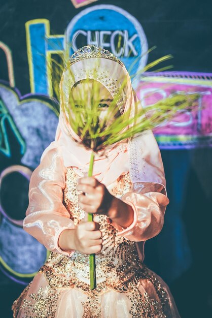 Foto niña con ropa tradicional sosteniendo una planta mientras está de pie contra la pared