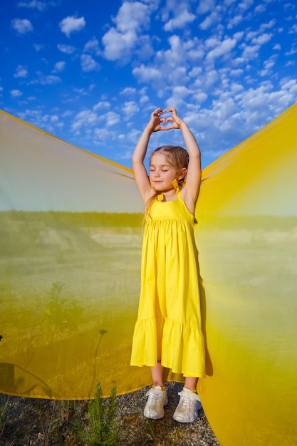 Una niña rubia de 7-8 años con un vestido amarillo brillante contra un cielo azul.