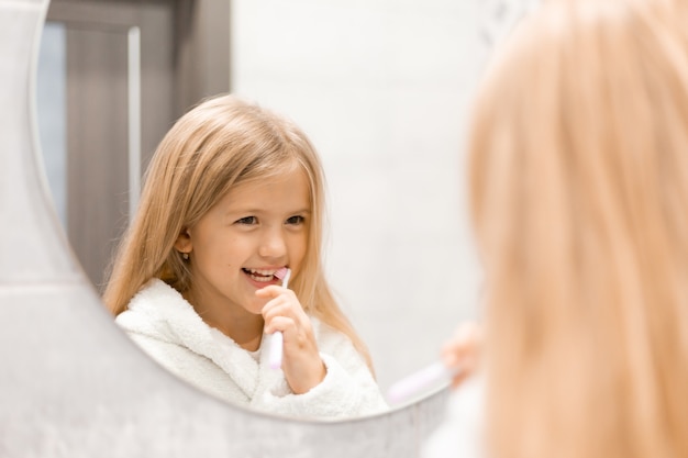 Niña rubia en una bata de baño blanca se cepilla los dientes frente al espejo del baño