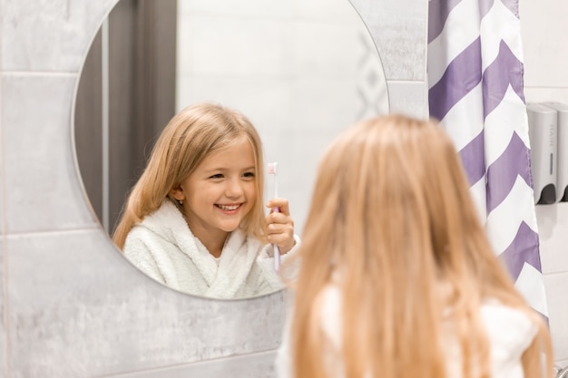 Niña rubia en una bata de baño blanca se cepilla los dientes frente al espejo del baño