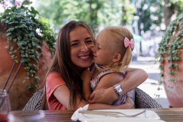 Foto niña rubia besa a mamá esperando un pedido en una cafetería.