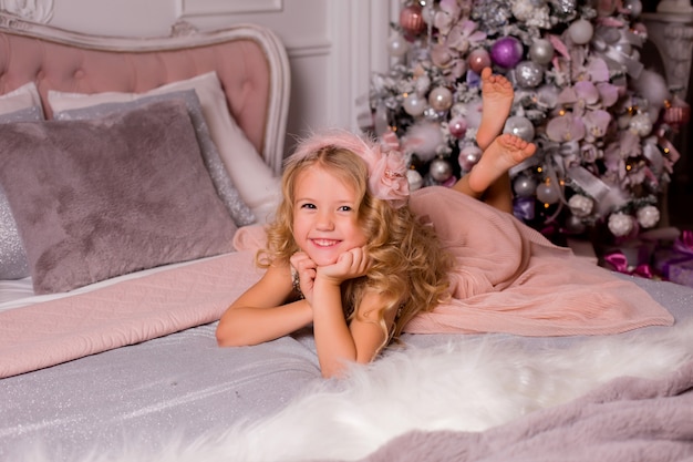 Niña rubia en la cama junto al árbol de Navidad. Nochebuena