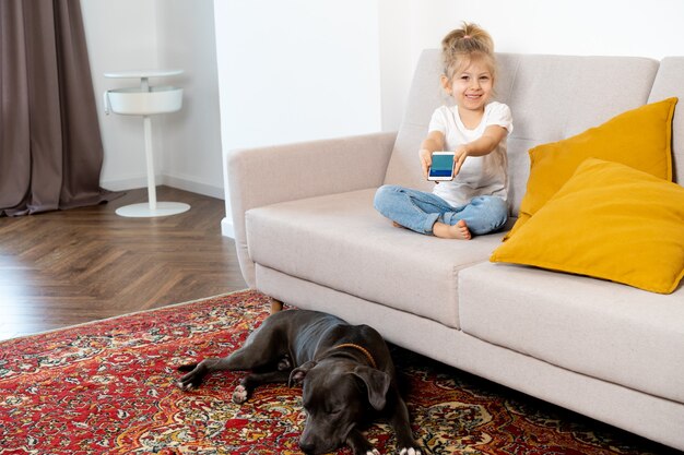 Una niña rubia en casa en el sofá con un teléfono móvil y un perro grande, niños y gadgets.