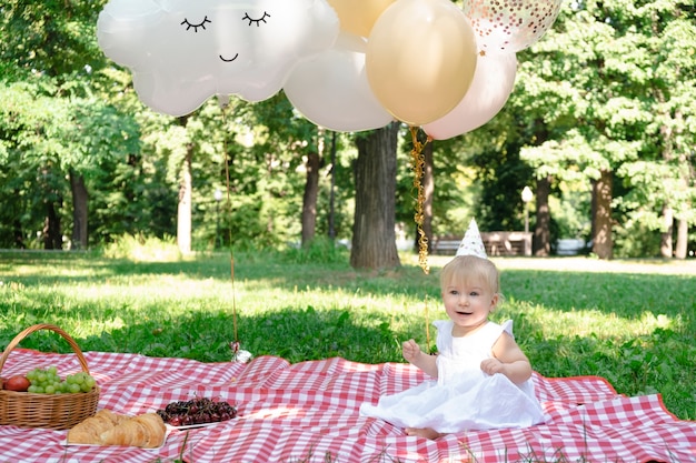 Niña rubia caucásica sonriendo celebrando el primer cumpleaños al aire libre en un picnic en el parque de verano