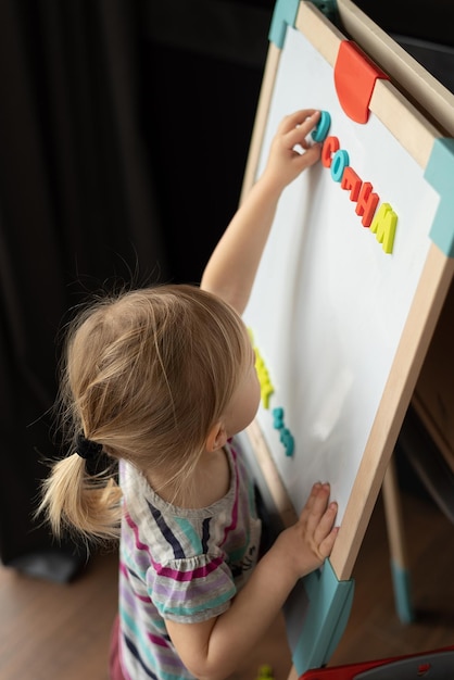 Foto una niña rubia se para cerca de un tablero magnético en un caballete para niños en la habitación
