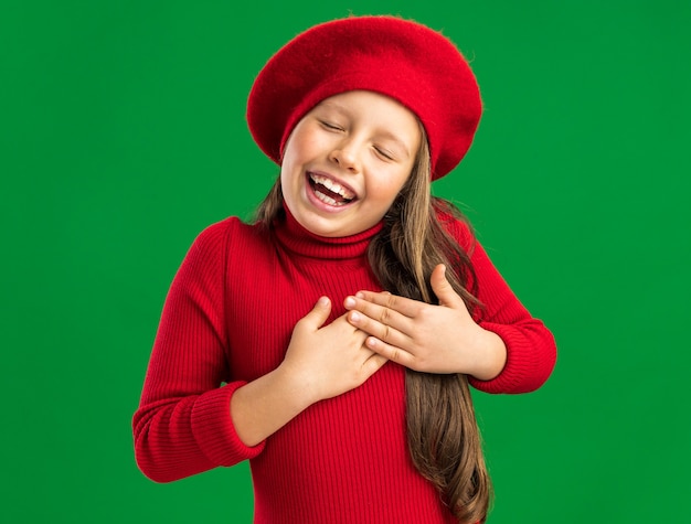 Niña rubia complacida con boina roja manteniendo las manos en el corazón con los ojos cerrados aislado en la pared verde con espacio de copia