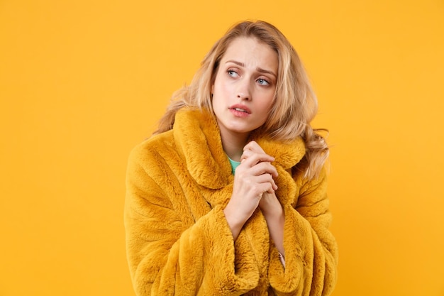 Niña rubia congelada con abrigo de piel amarillo posando aislada en el fondo de la pared naranja, retrato de estudio. Gente emociones sinceras concepto de estilo de vida. Simulacros de espacio de copia. Frotando las manos calientes.