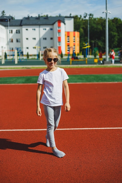 Una niña rubia corre en el campo de deportes y sonríe, lleva gafas de sol y una camiseta blanca Vacaciones de verano y tiempo de vacaciones