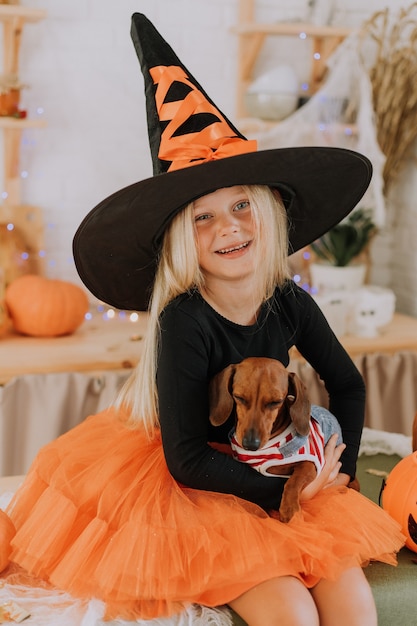 Niña rubia con un disfraz de bruja con un enorme sombrero de bruja y una falda hinchada naranja sostiene un perro salchicha