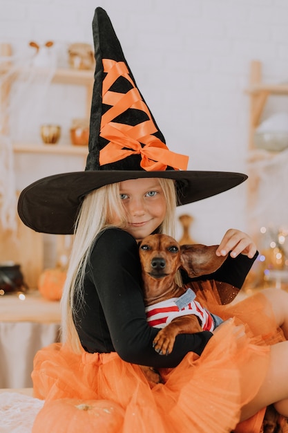 Niña rubia con un disfraz de bruja con sombrero de bruja y una falda hinchada naranja sostiene un perro salchicha enano