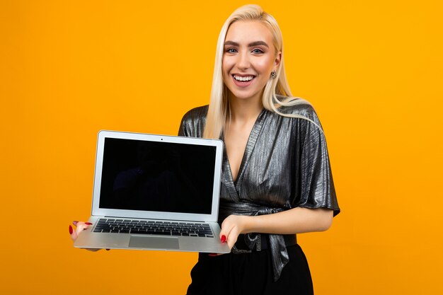 Niña rubia encantadora sonriente sosteniendo una pantalla de portátil en blanco en un espacio de pared amarilla