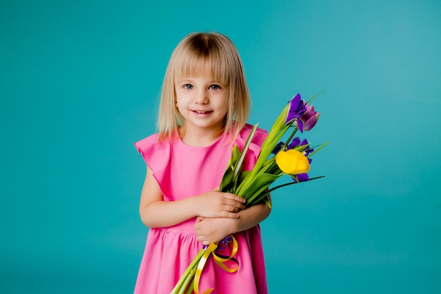 Niña rubia está sonriendo con un vestido rosa y sosteniendo un ramo de flores de primavera en un espacio azul aislado