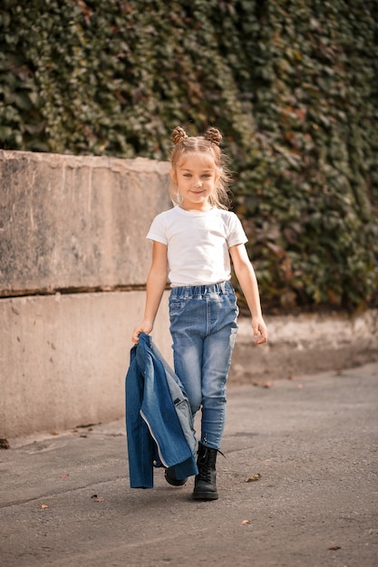 Niña rubia con estilo en jeans y una camiseta blanca camina por la calle. Niña de 7 años modelo pequeña, hermosa niña