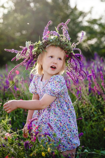 Una niña rubia feliz con una corona de salvia y un vestido colorido en un campo floreciente de verano