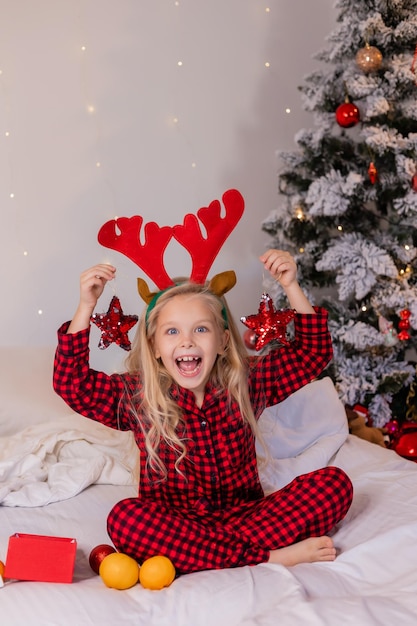 una niña rubia feliz en pijama en Navidad mira los regalos de Año Nuevo y juega con Año Nuevo