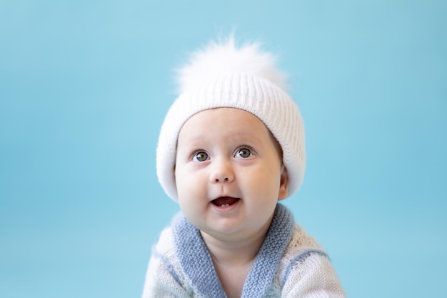 Una niña rubia con un gorro de punto de invierno blanco y un suéter de punto sobre un fondo azul aísla el espacio para texto bebé niño pequeño con ropa de invierno