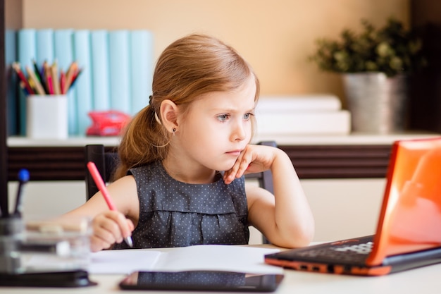 Niña rubia haciendo los deberes en casa en la mesa. El niño es educado en casa. Una chica con cabello claro realiza una tarea en línea usando una computadora portátil y tableta.