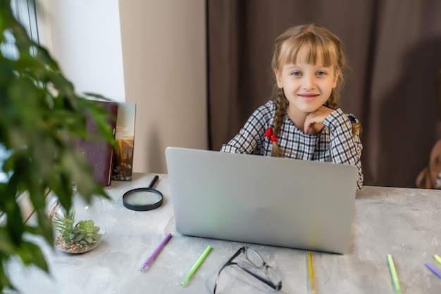 Niña rubia haciendo los deberes en casa en la mesa. El niño es educado en casa. Una niña de cabello claro realiza una tarea en línea usando una computadora portátil y una tableta.