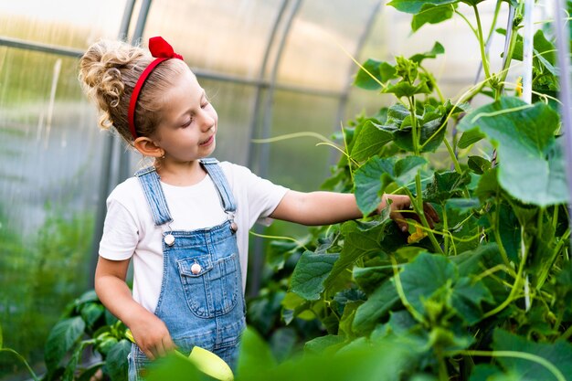 Una niña rubia en un invernadero con un omóplato y un rastrillo se encarga de las plantas
