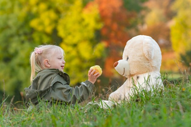 Niña rubia juega con un gran oso de peluche sentado en la hierba en el fondo del bosque de otoño