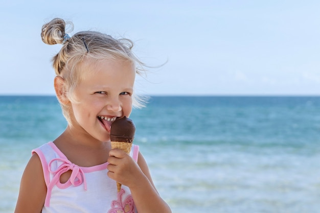 Niña rubia lame helado de chocolate y sonríe sobre un fondo de cielo y mar